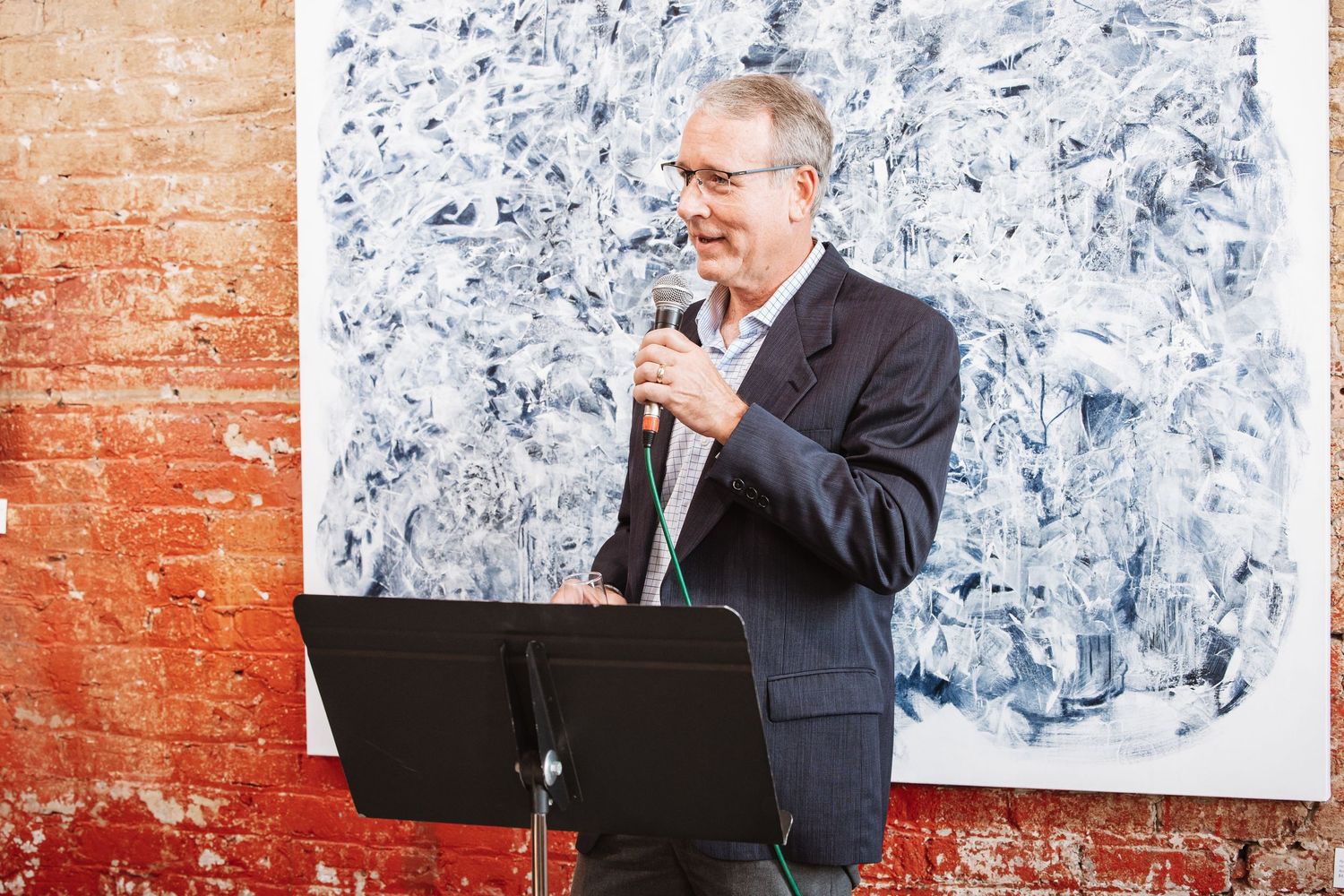 Man in blazer speaking into microphone at podium in front of abstract artwork in brick gallery.