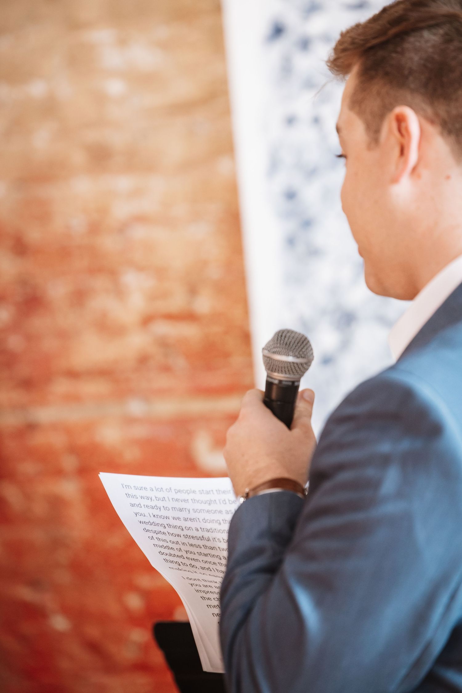 Over shoulder view of man in blue suit holding microphone and reading speech at wedding.