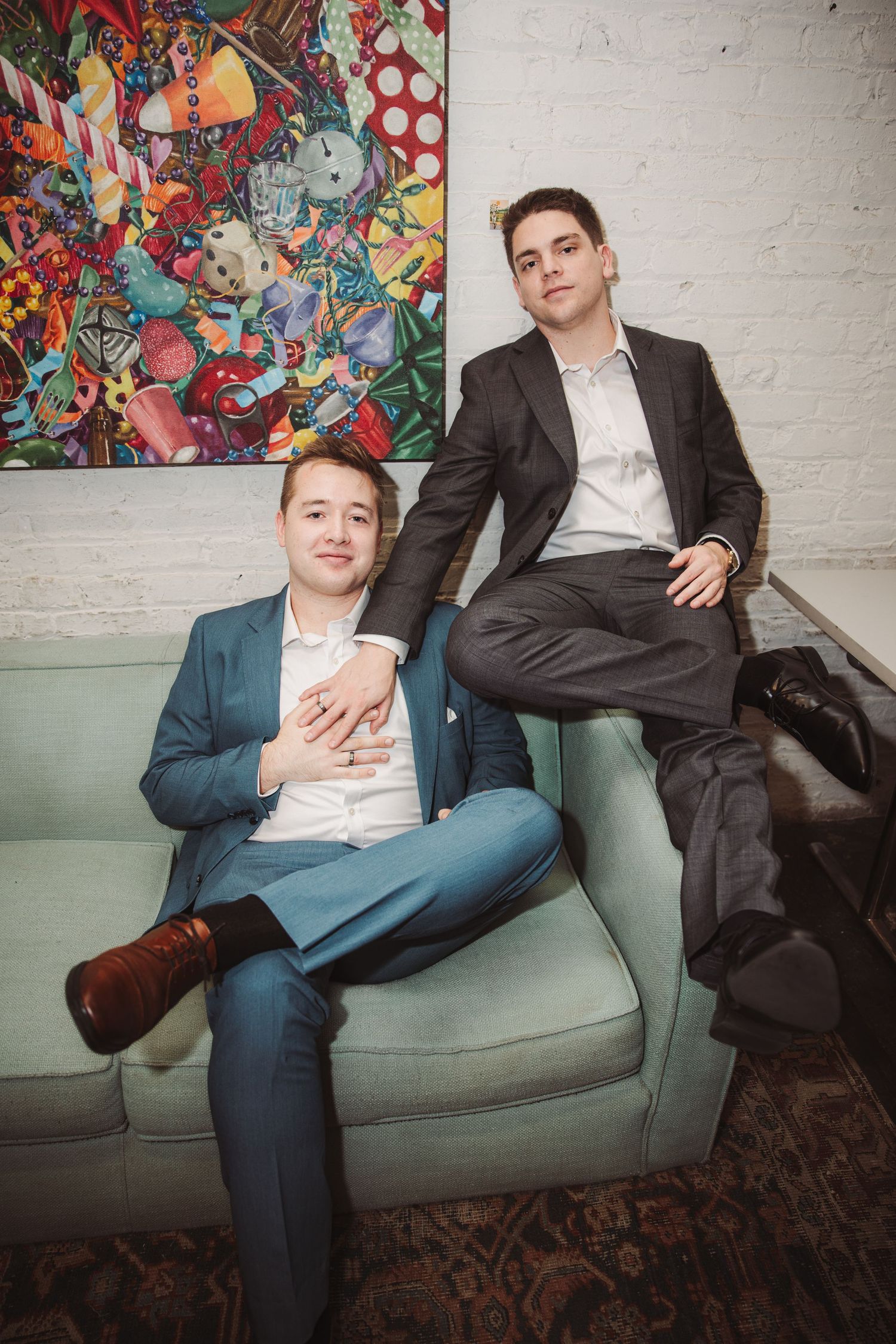 Two young men in suits pose on a teal sofa in front of vibrant colorful artwork in a modern space.