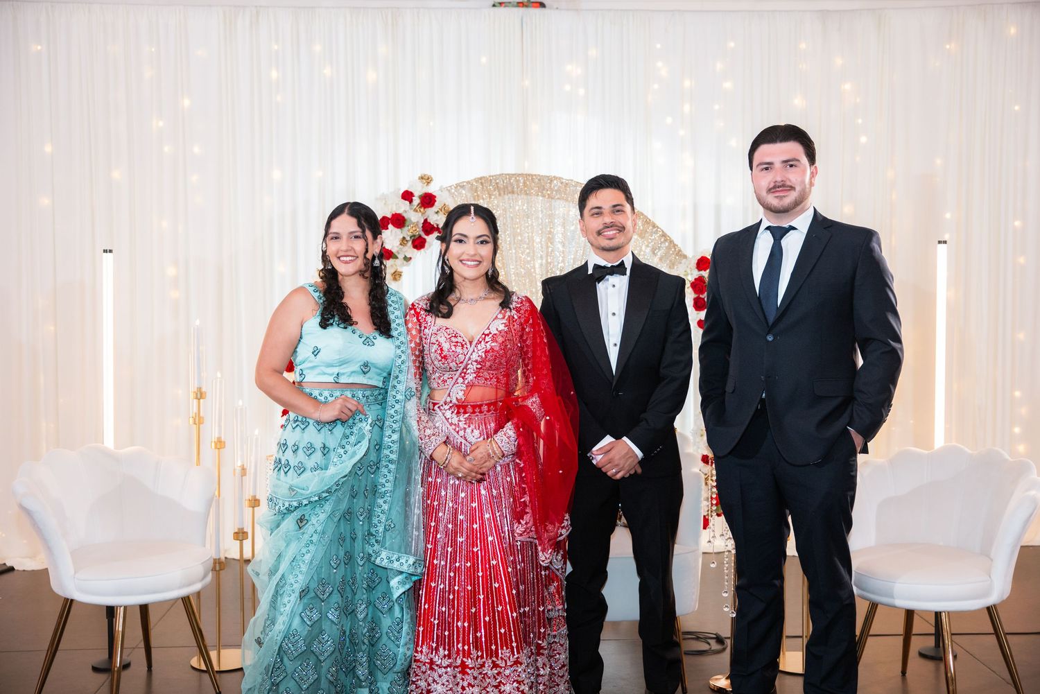 Bride in red lehenga and groom in tuxedo pose with two guests at a decorated wedding venue.