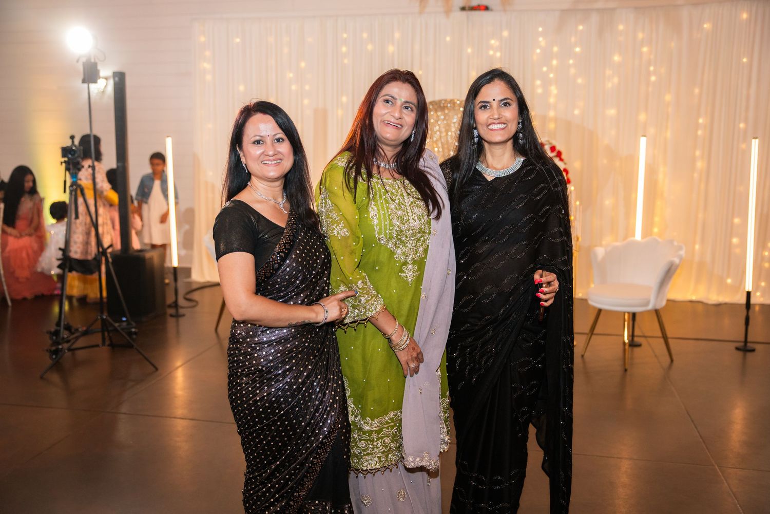 Three women in elegant South Asian attire pose together at a formal event with fairy lights backdrop.