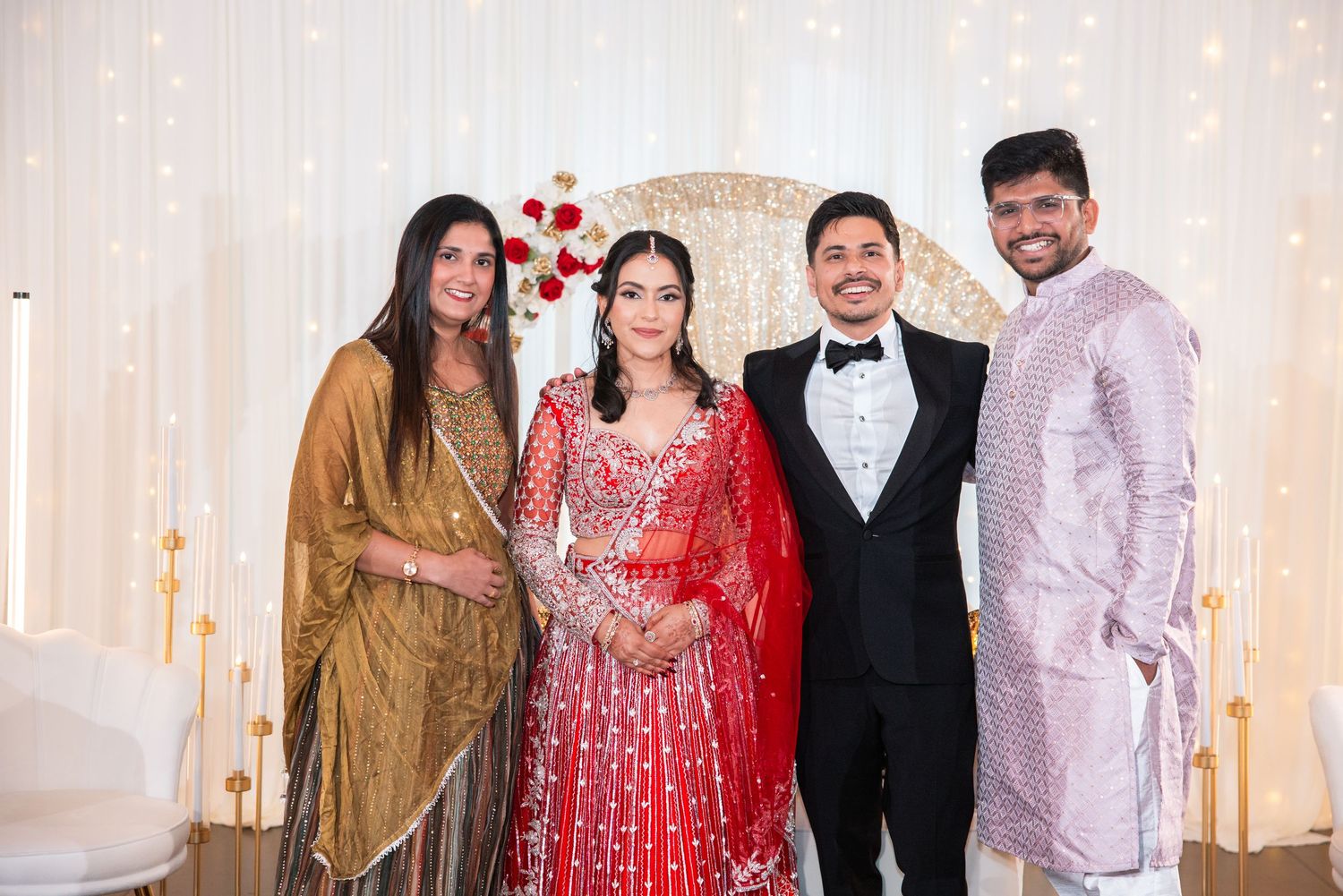 Four people in Indian wedding attire smiling together at decorated reception with floral backdrop.
