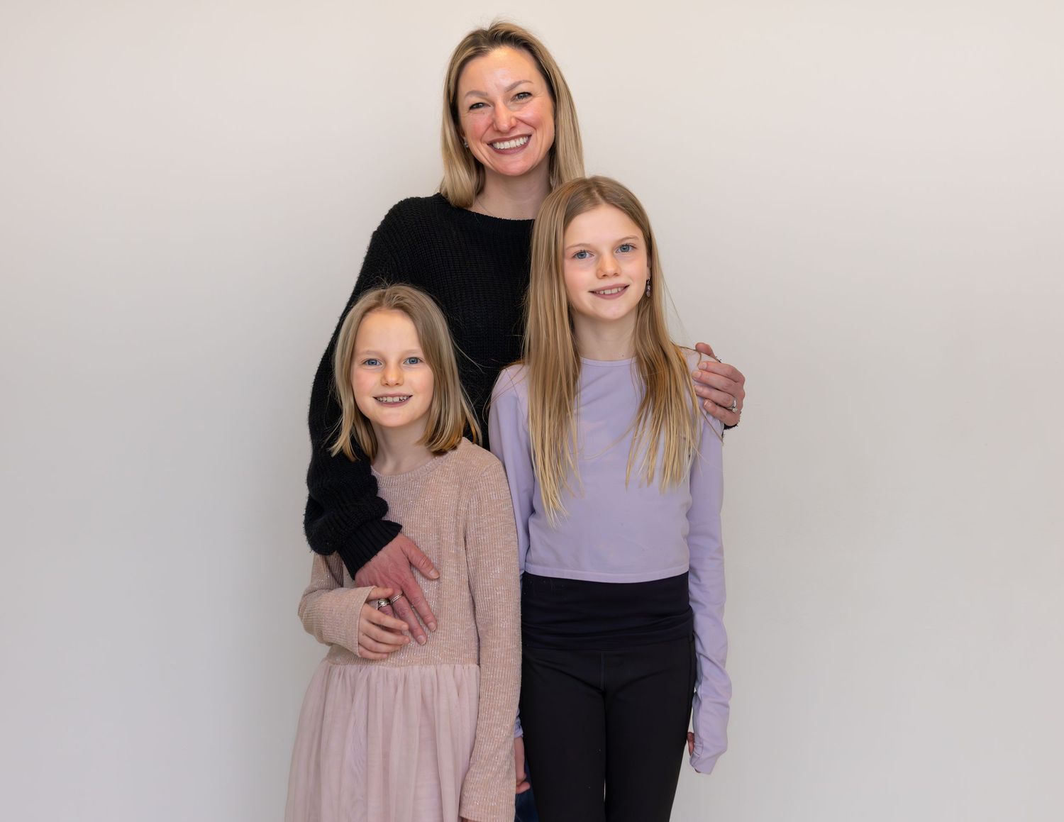 A smiling mother poses with her two blonde daughters in a bright studio portrait.