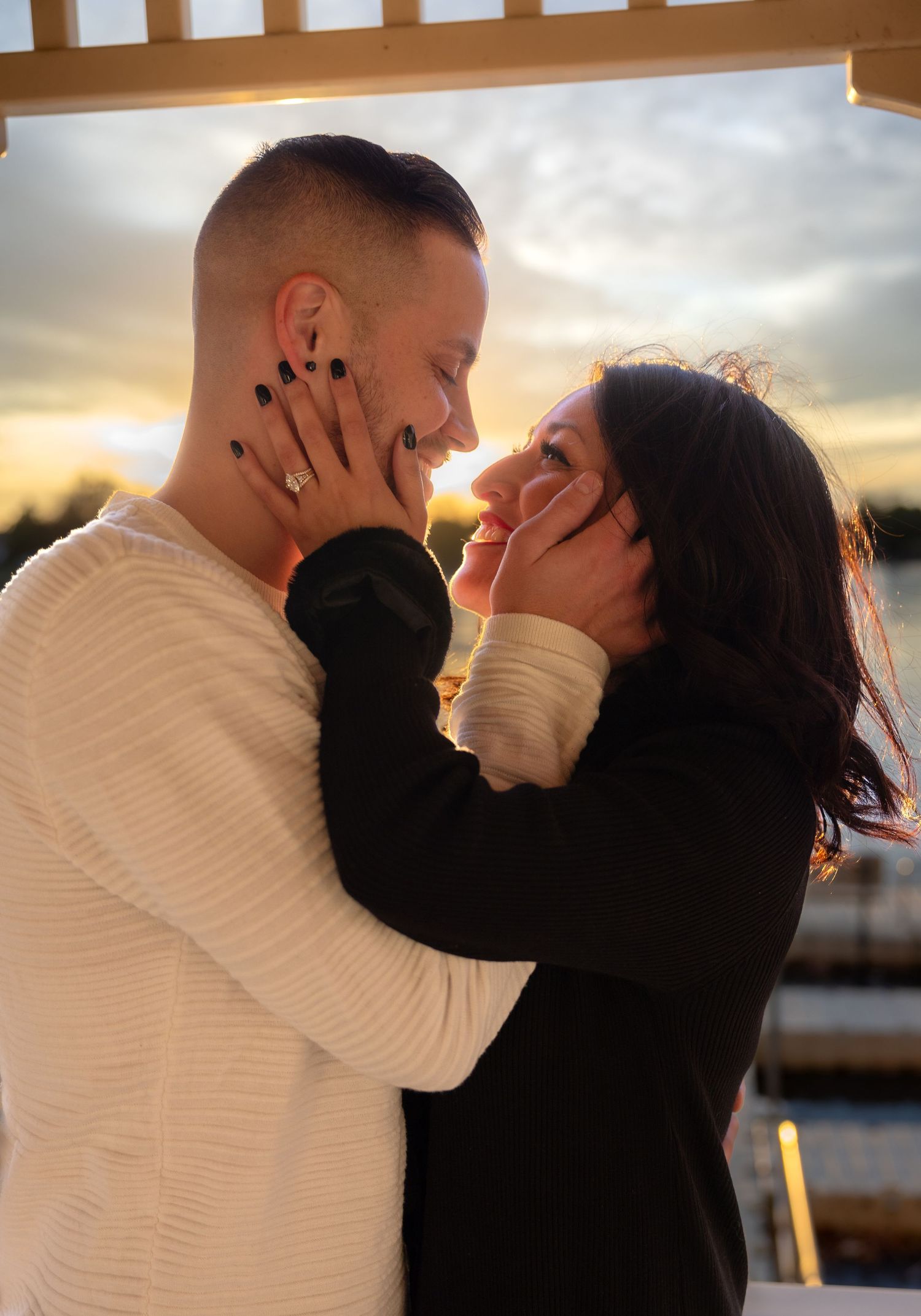 Couple nearly kissing at golden hour sunset, woman with black nails cradling mans face outdoors.
