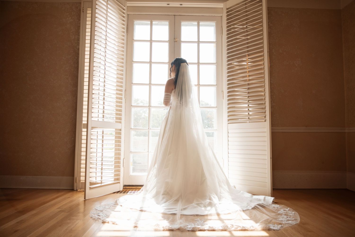Bride in elegant white gown with cathedral veil standing by sunlit French doors in a warmly lit room.