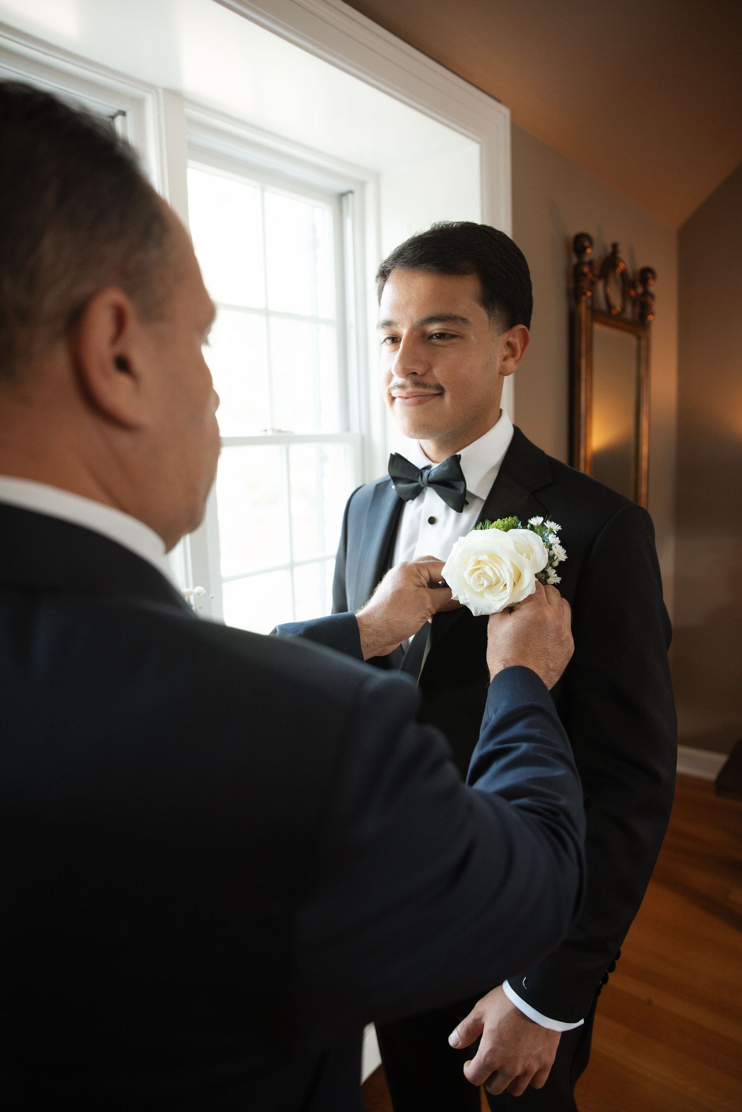 Older man pinning white rose boutonniere on smiling young groom in black tuxedo near window.