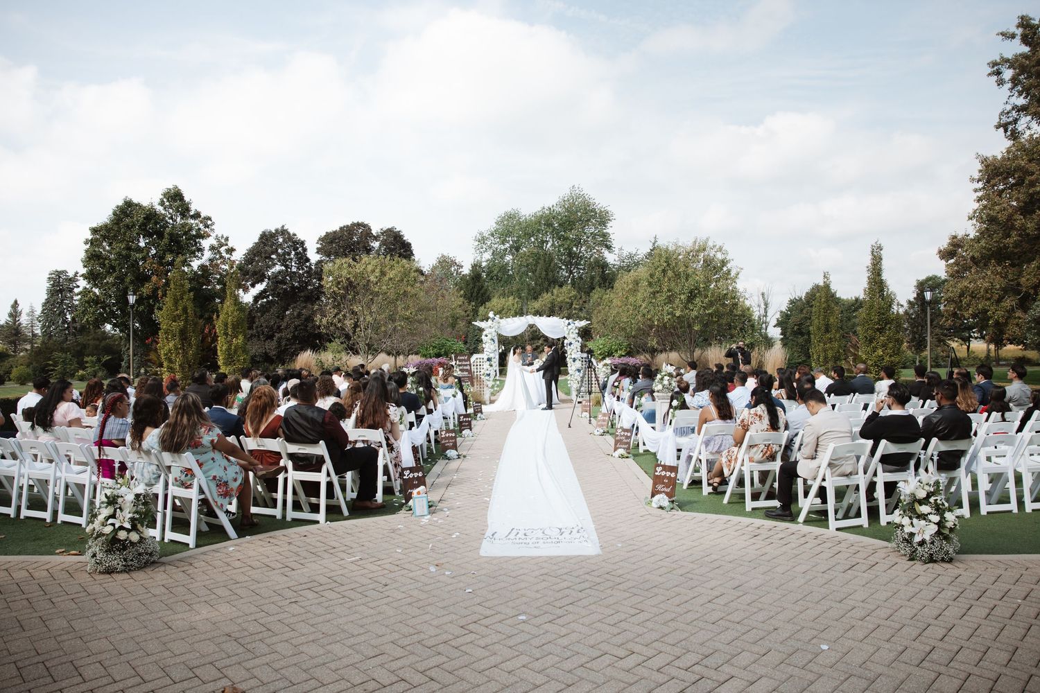 Outdoor wedding ceremony with white chairs, floral arch, and long aisle runner.