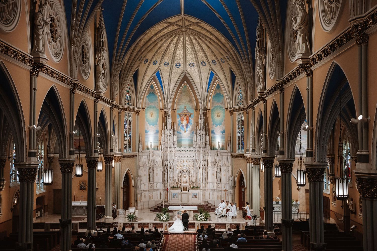 Wide view of stunning Gothic Catholic church interior during wedding ceremony with vaulted blue ceiling.
