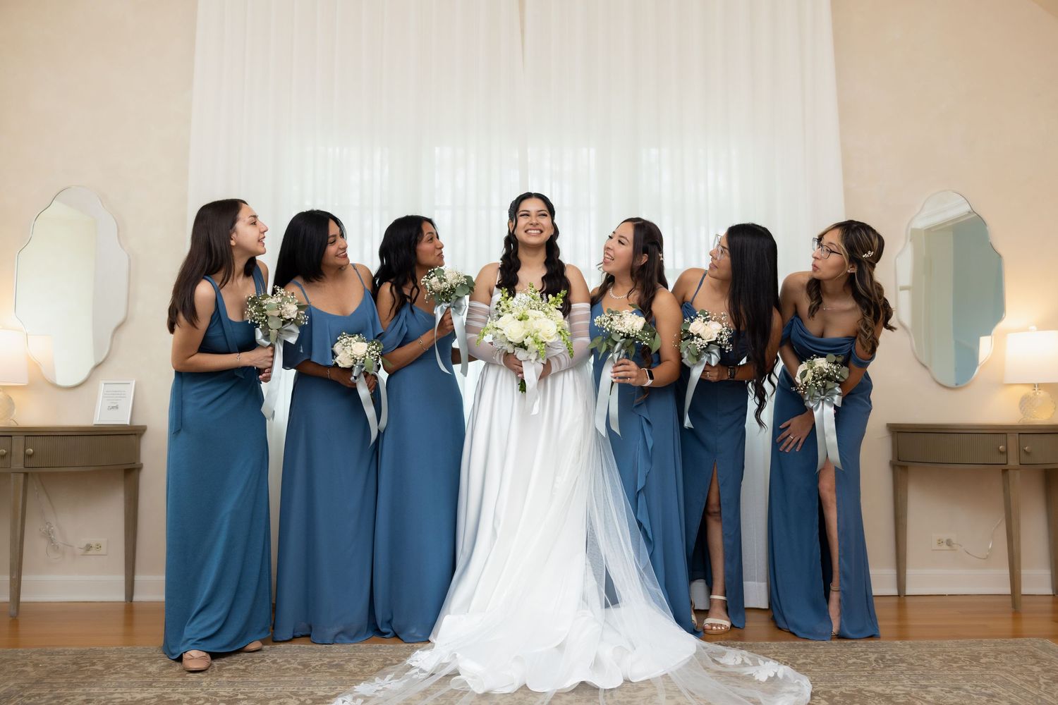Bride in white gown poses with six bridesmaids in steel blue dresses holding bouquets indoors.