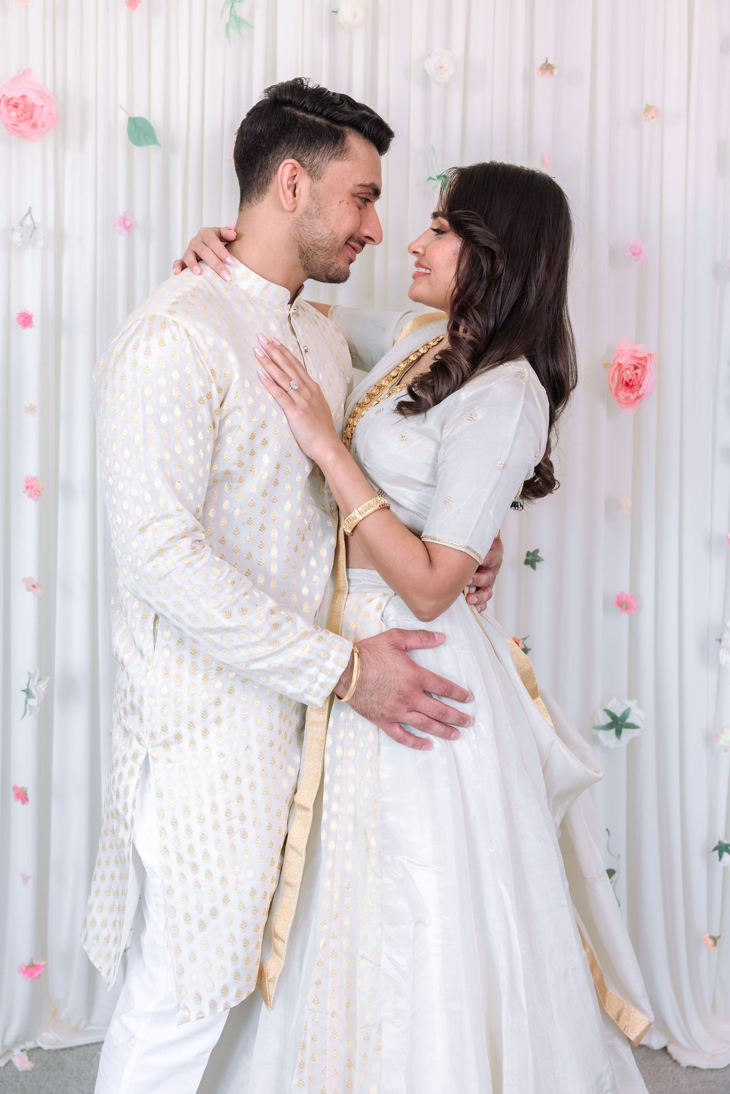 Indian couple in matching white and gold traditional outfits sharing a romantic moment at their engagement ceremony.