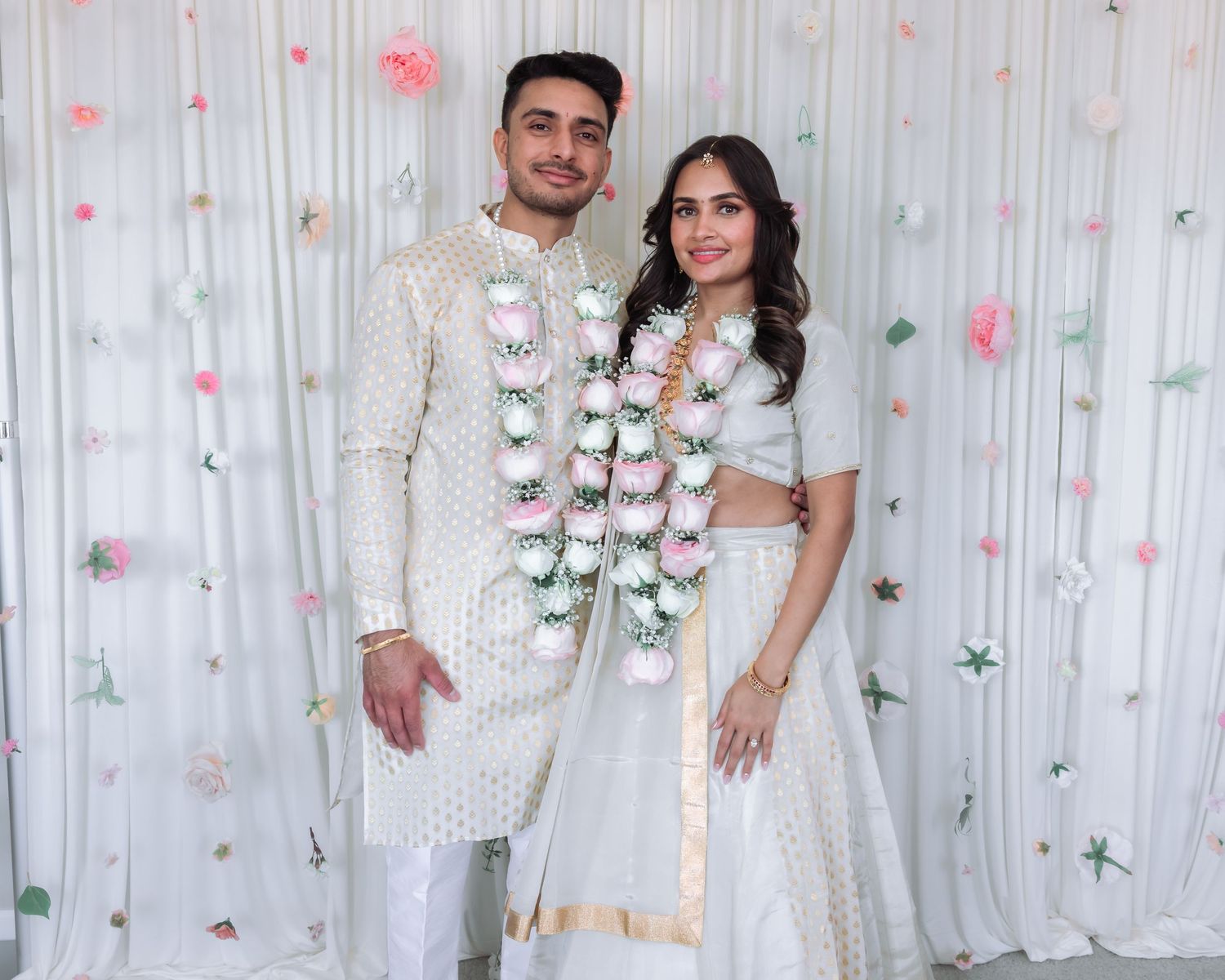Indian couple in elegant white traditional attire pose with floral garlands at engagement ceremony.