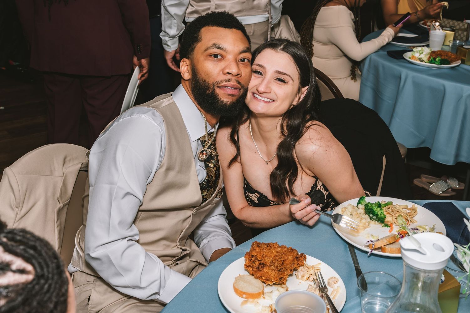 A couple poses happily at a wedding reception dinner table with plates of food in front of them.