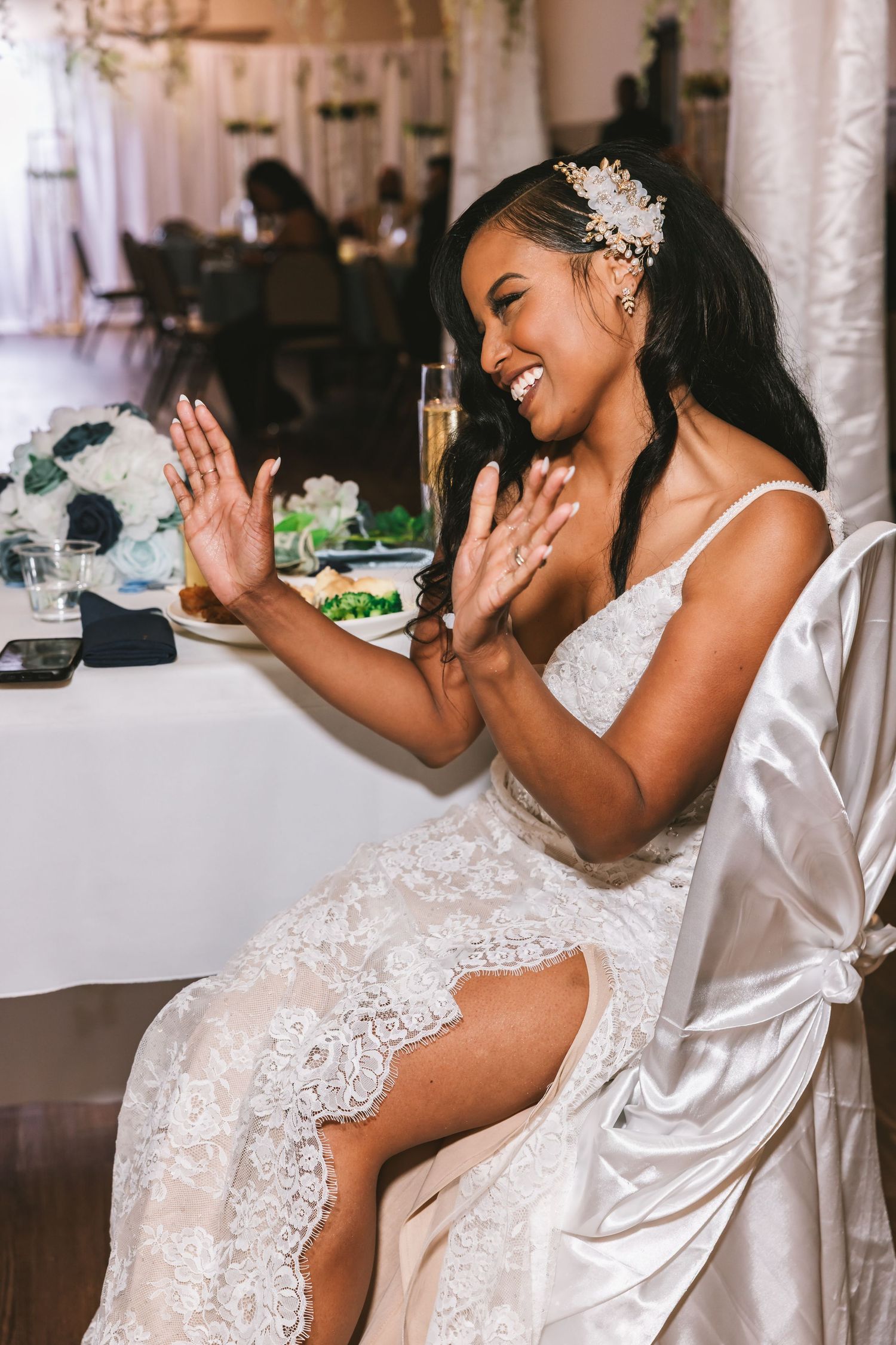 Joyful bride in lace gown claps and laughs at wedding reception table with champagne.