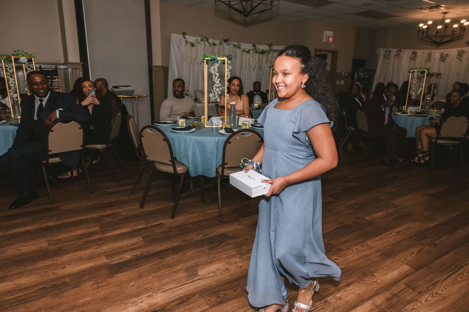 Young girl in blue dress carries a gift box across the dance floor at a wedding reception.
