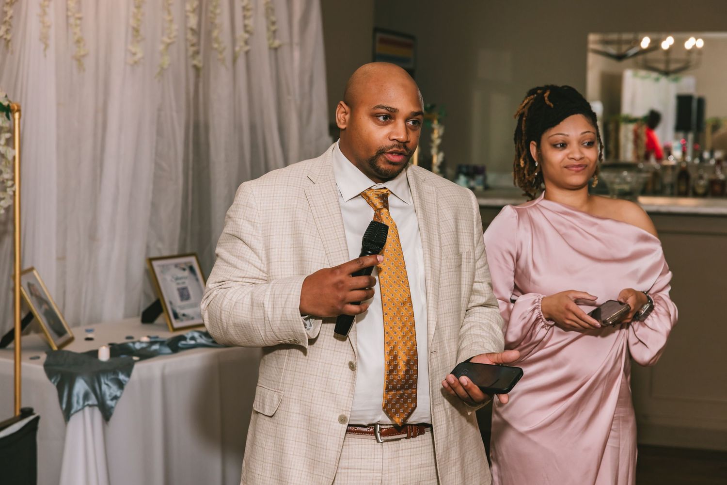 Man in beige suit giving speech at wedding reception while woman in pink dress stands nearby.