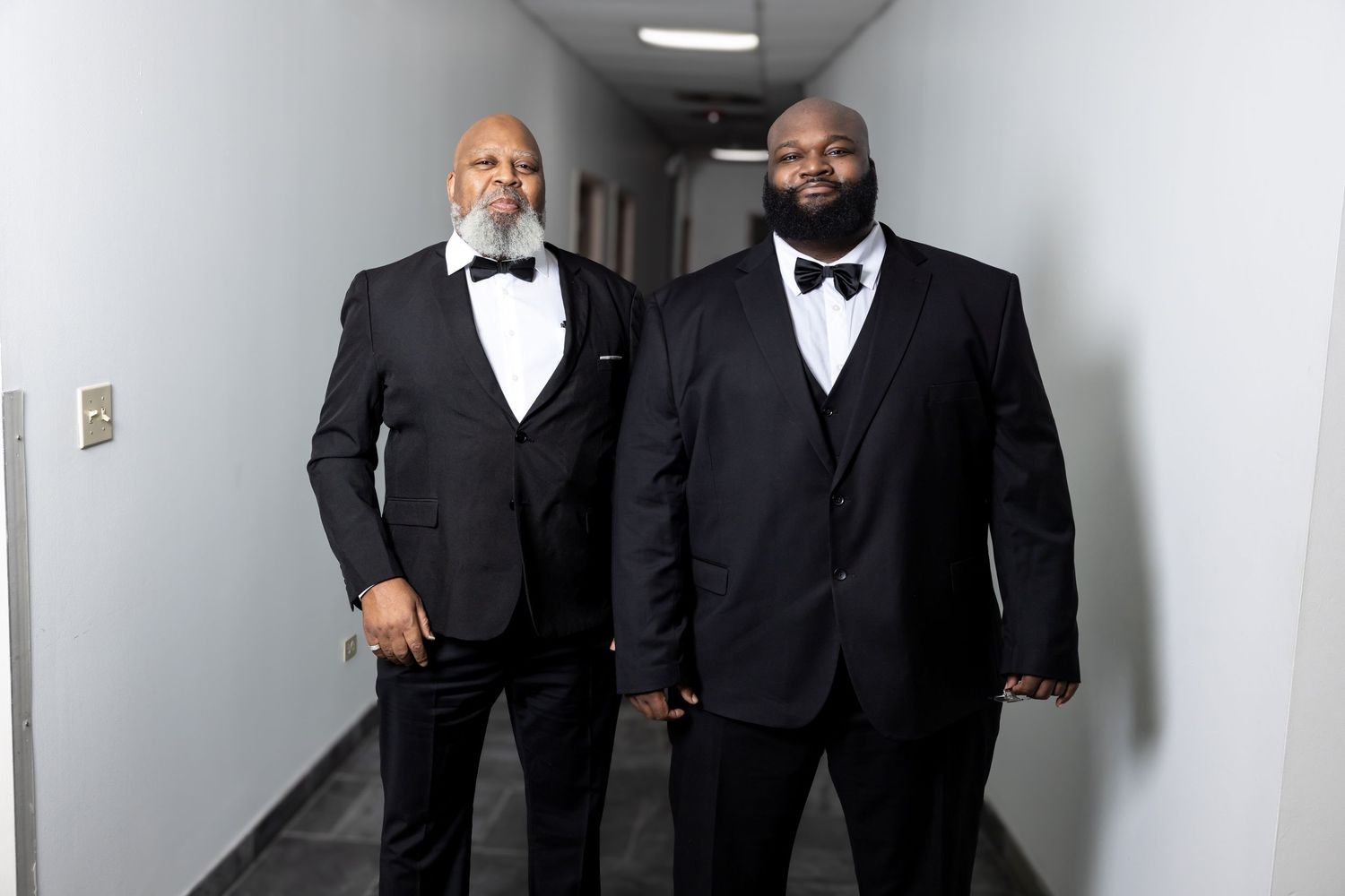 Two men in black tuxedos with bow ties walking confidently down a bright hallway together.