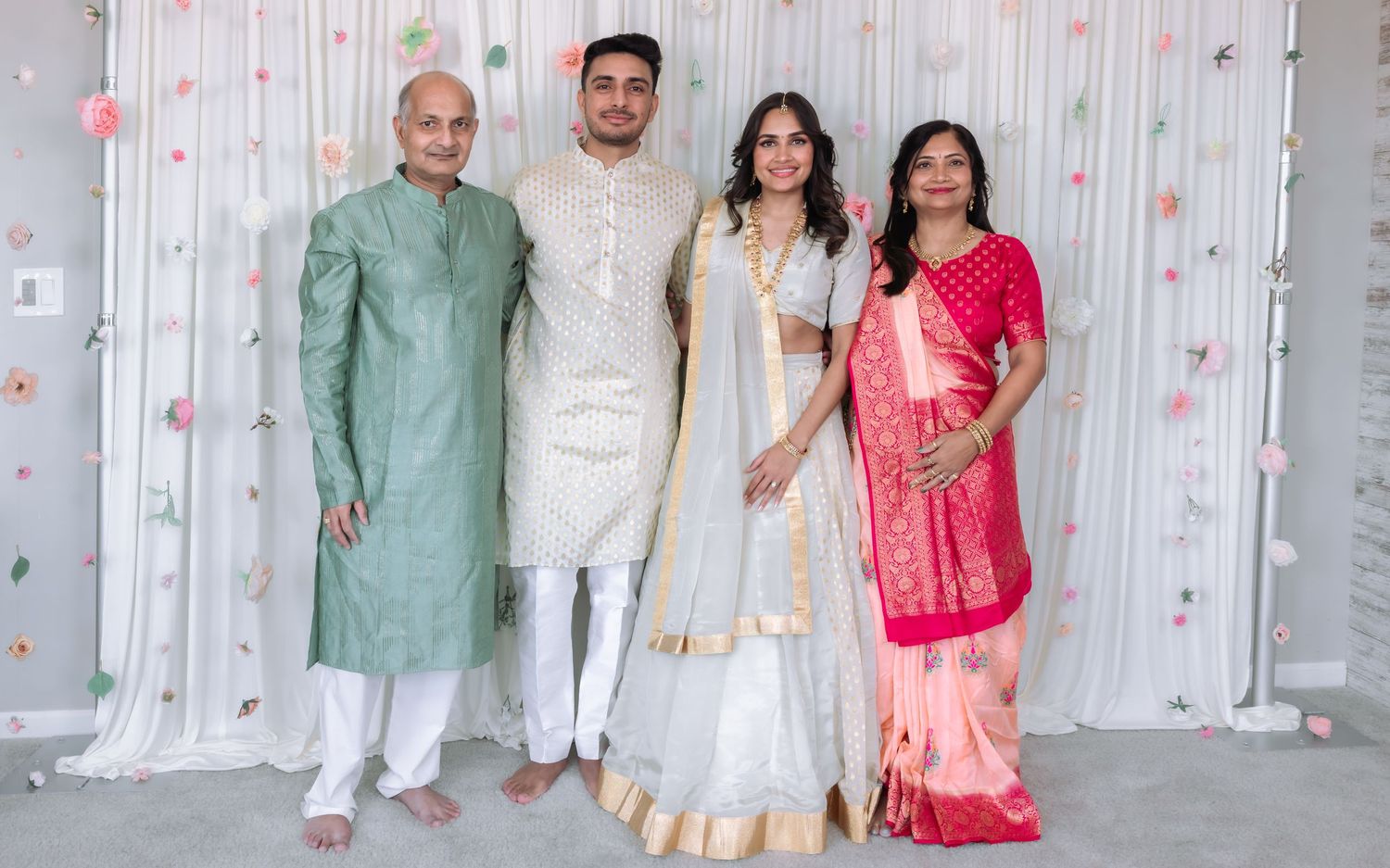 Indian wedding couple with garlands pose with family in traditional attire before floral backdrop.