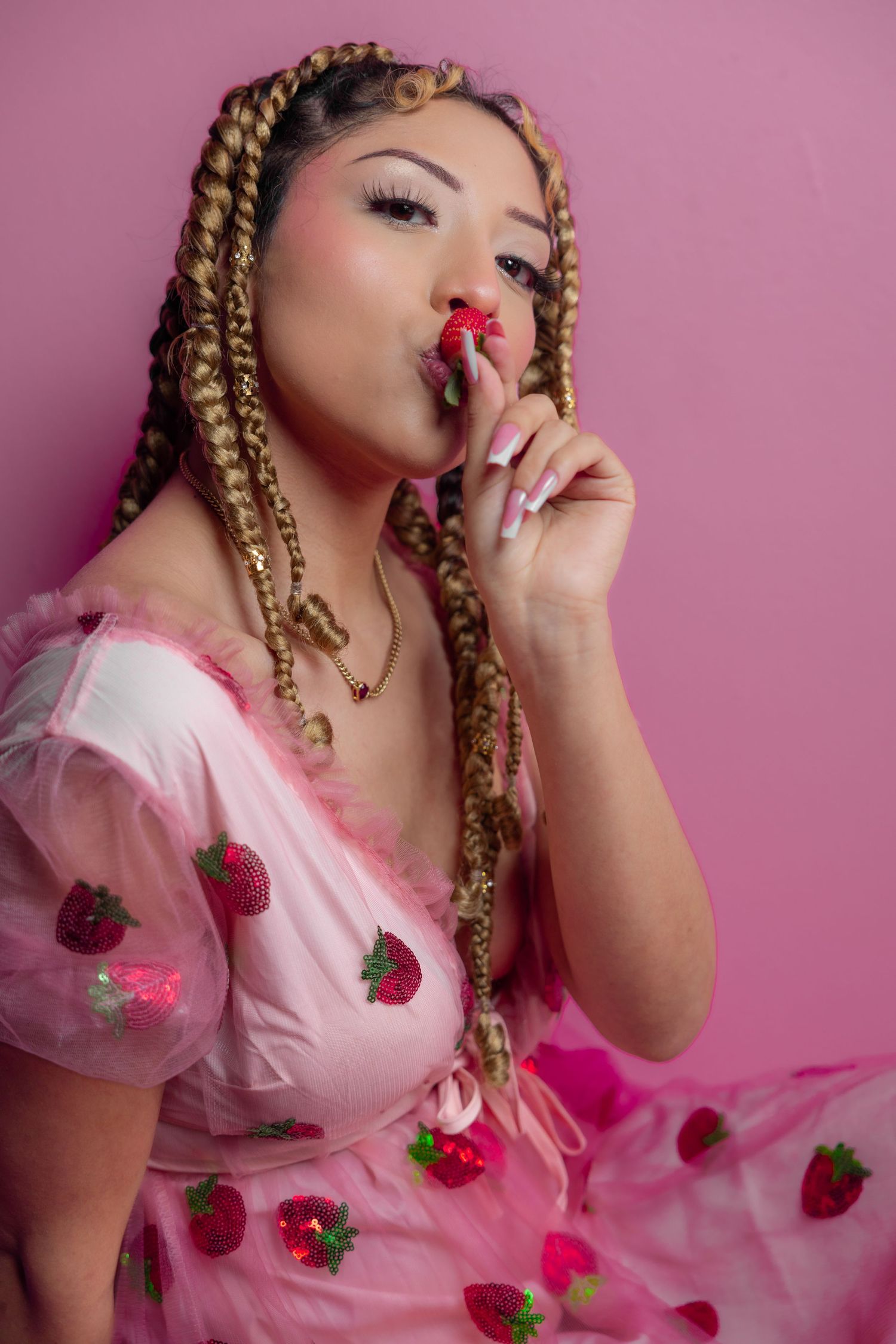 Woman in pink strawberry tulle dress eating a strawberry against pink background.
