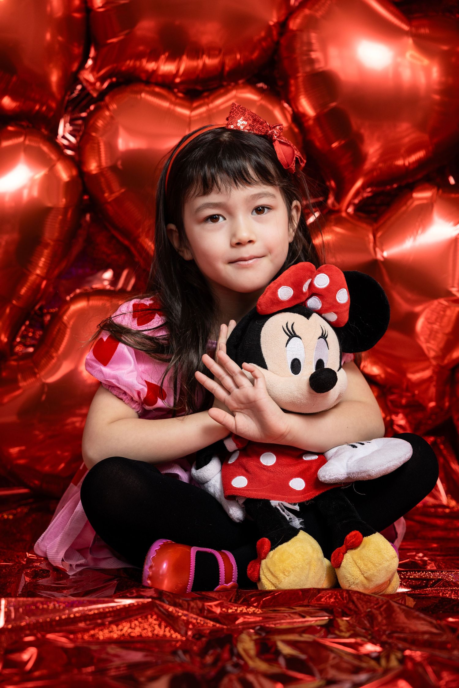 Young girl hugging Minnie Mouse plush toy surrounded by red heart-shaped balloons.