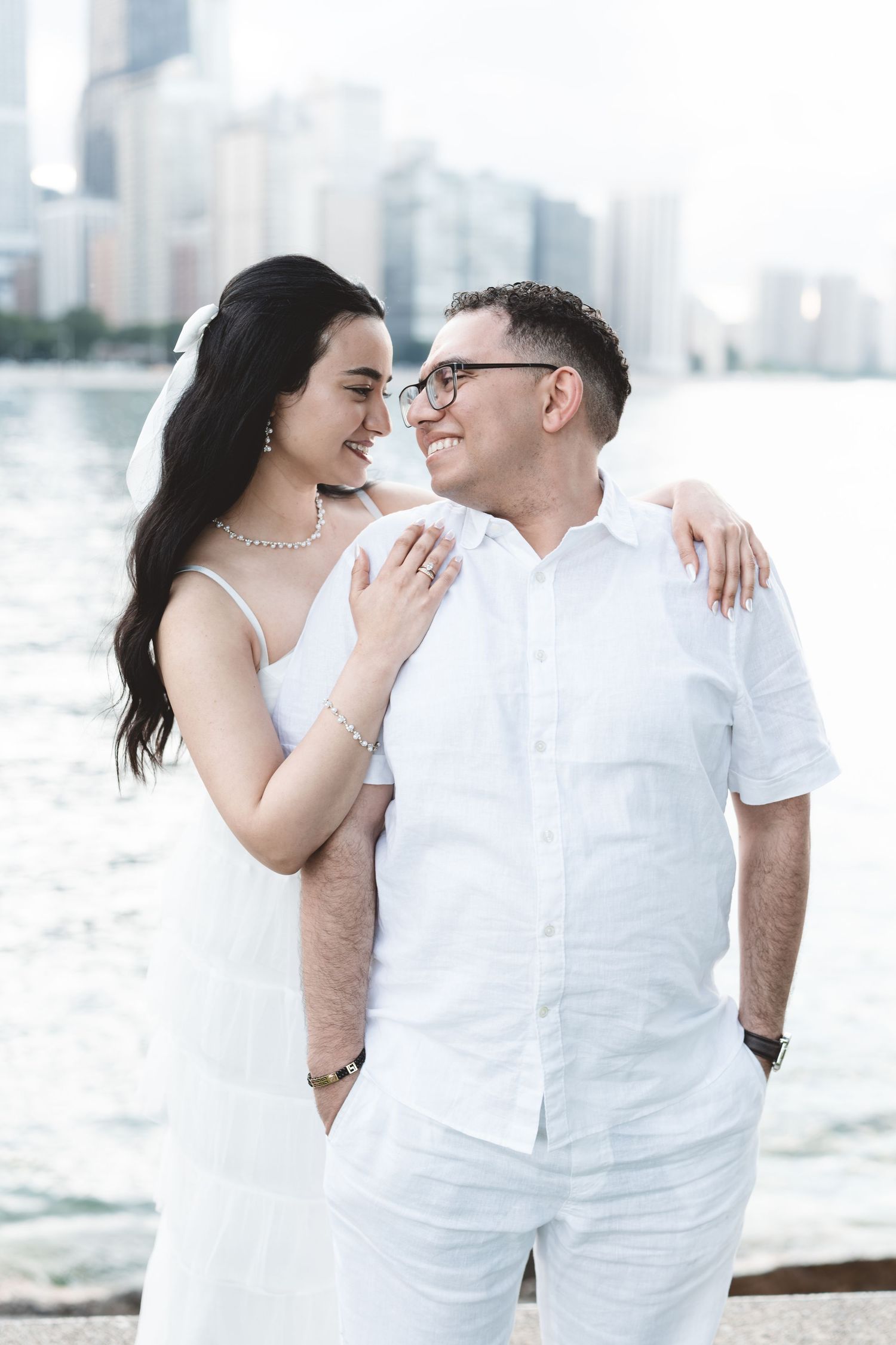 Couple in white attire sharing a romantic moment by the waterfront with city skyline backdrop.