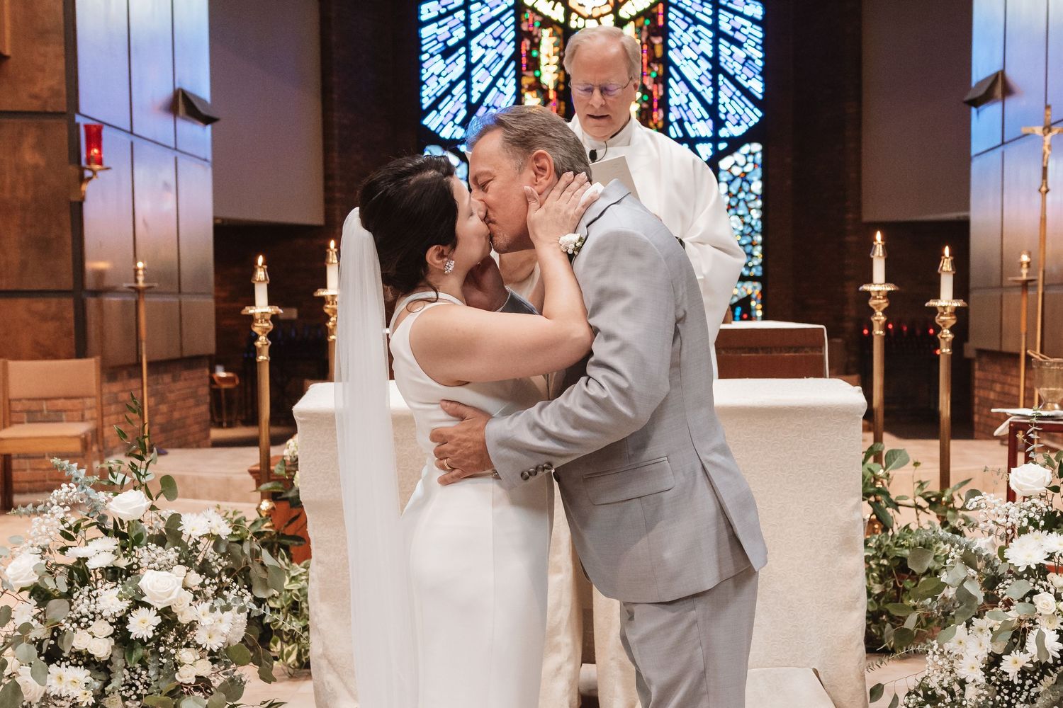 Bride and groom share first kiss at church altar surrounded by white floral arrangements.