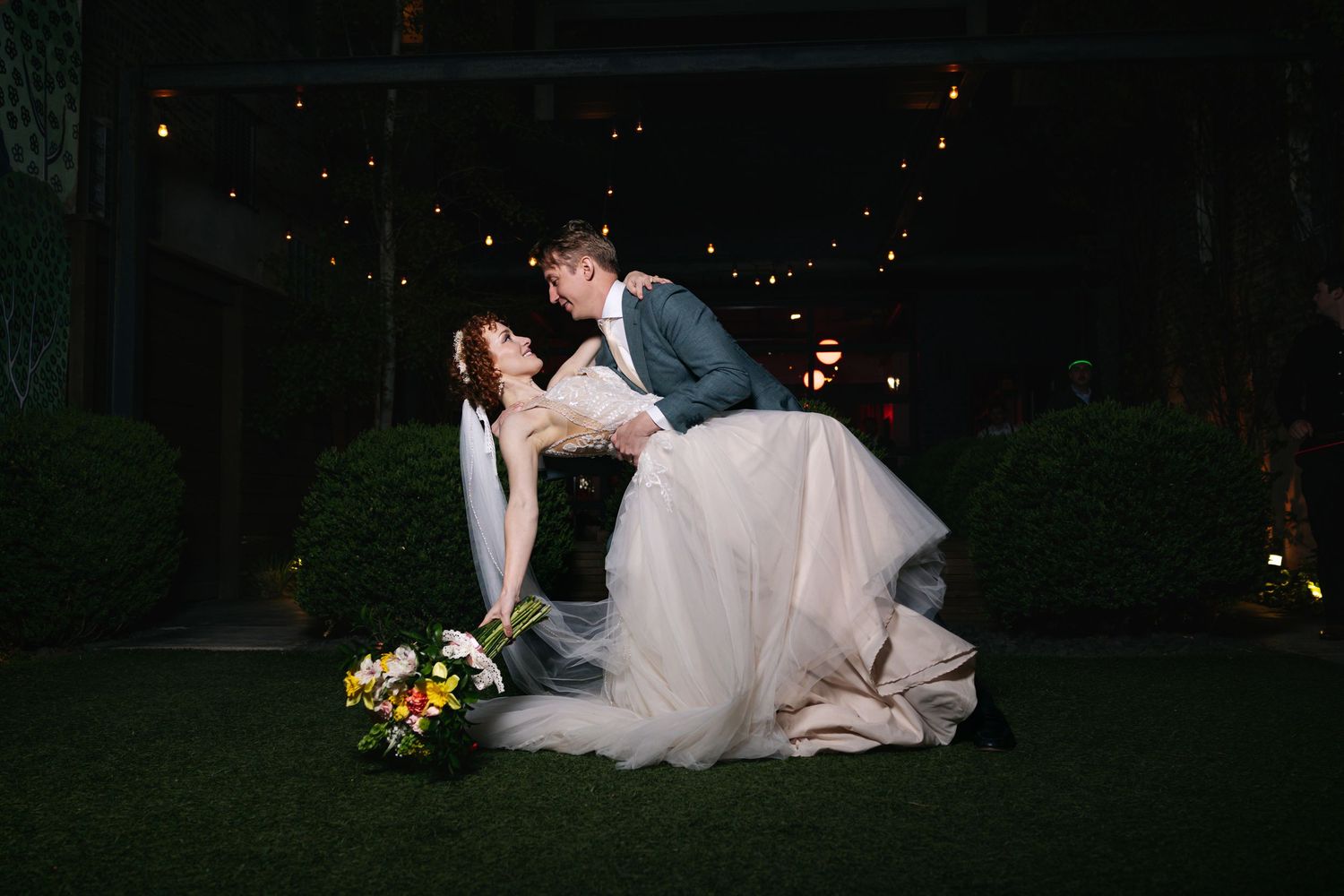 Groom dips bride in white tulle gown for romantic kiss under string lights on green lawn at night.