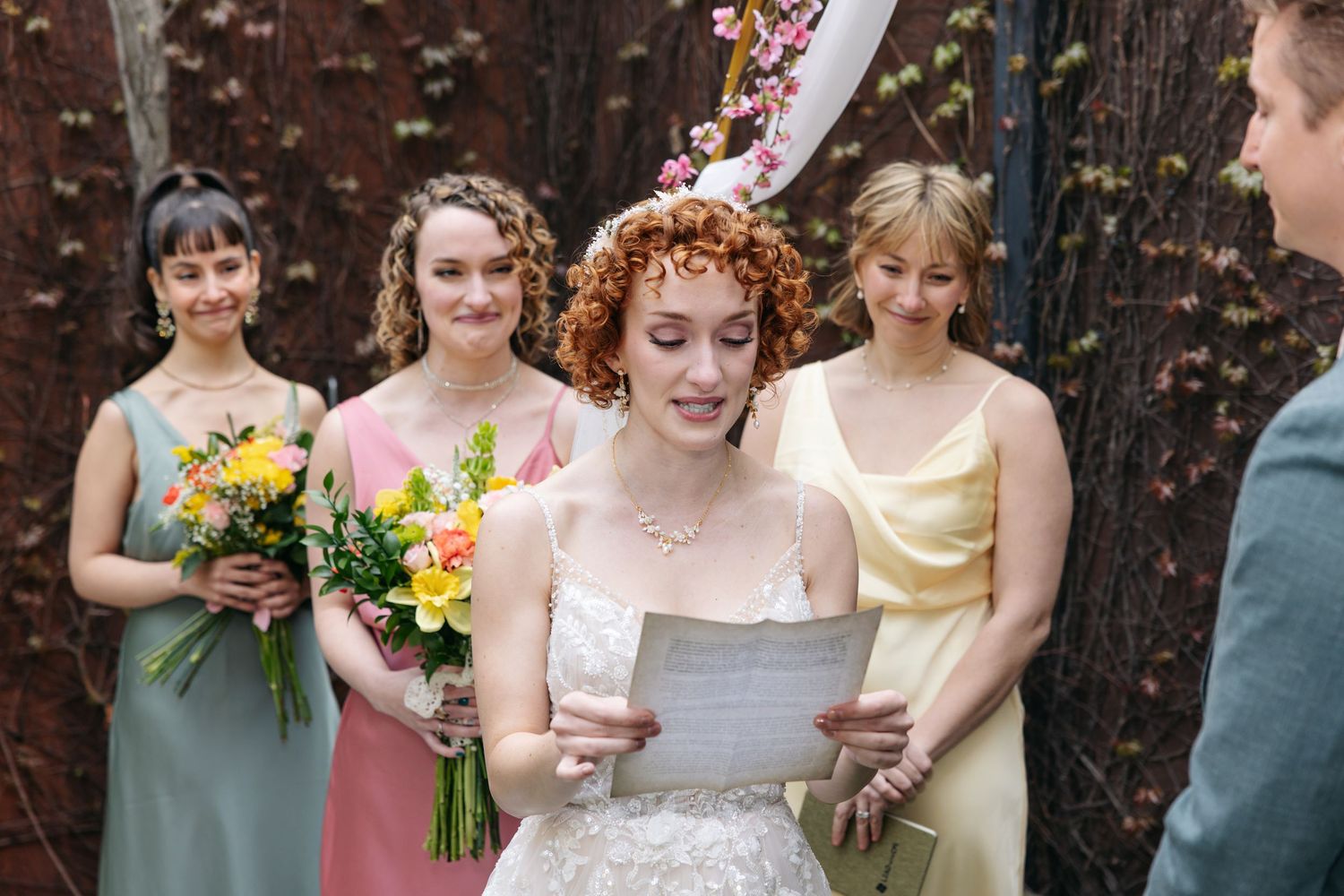 Bride reads vows at outdoor wedding ceremony with bridesmaids in colorful dresses holding yellow flowers.