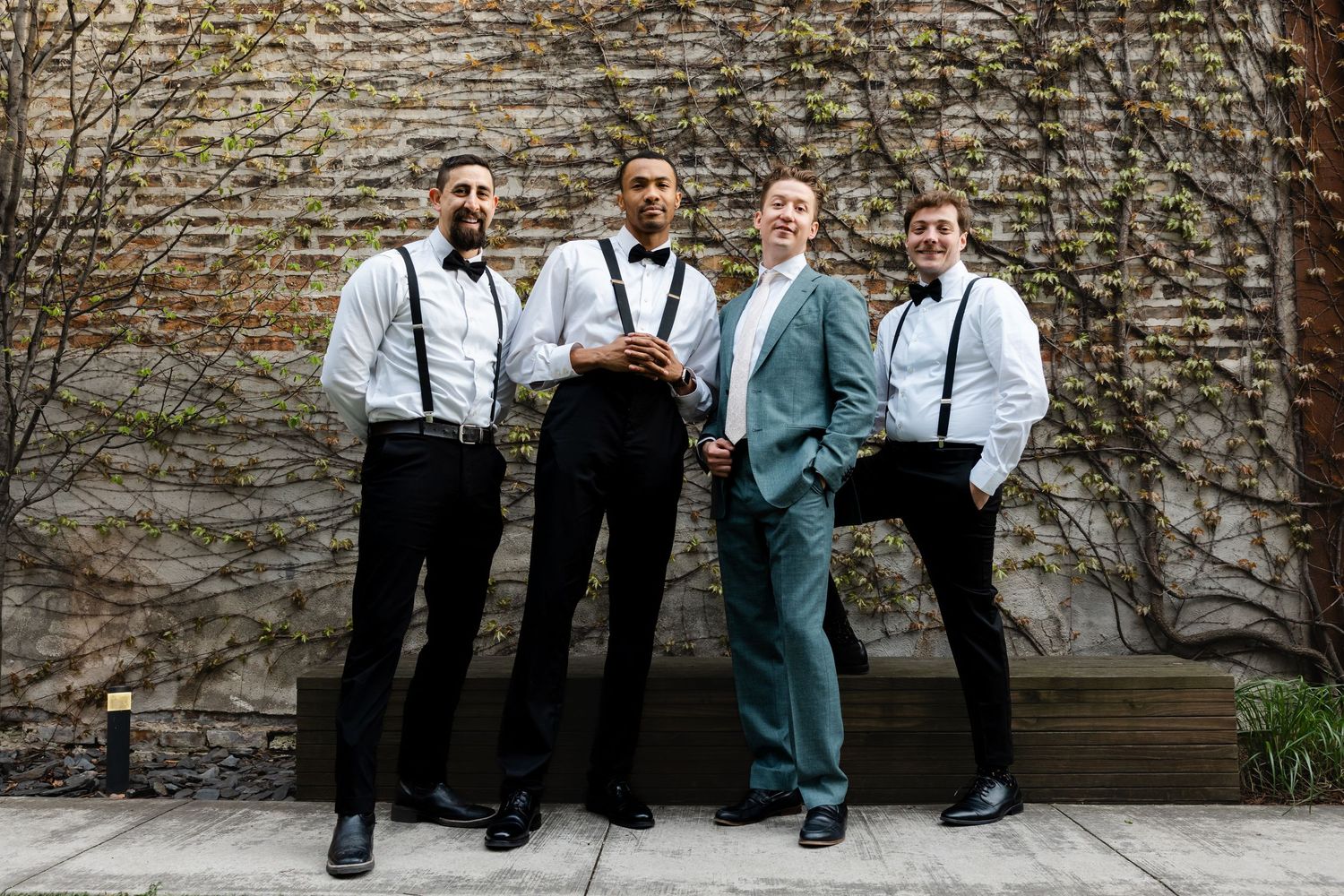 Groom in teal suit poses with three groomsmen in suspenders against ivy-covered brick wall.