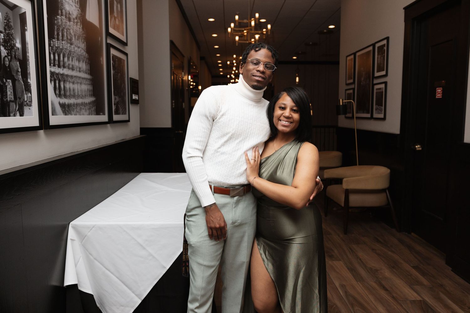 Couple poses together in a restaurant, man in white turtleneck and woman in green dress.