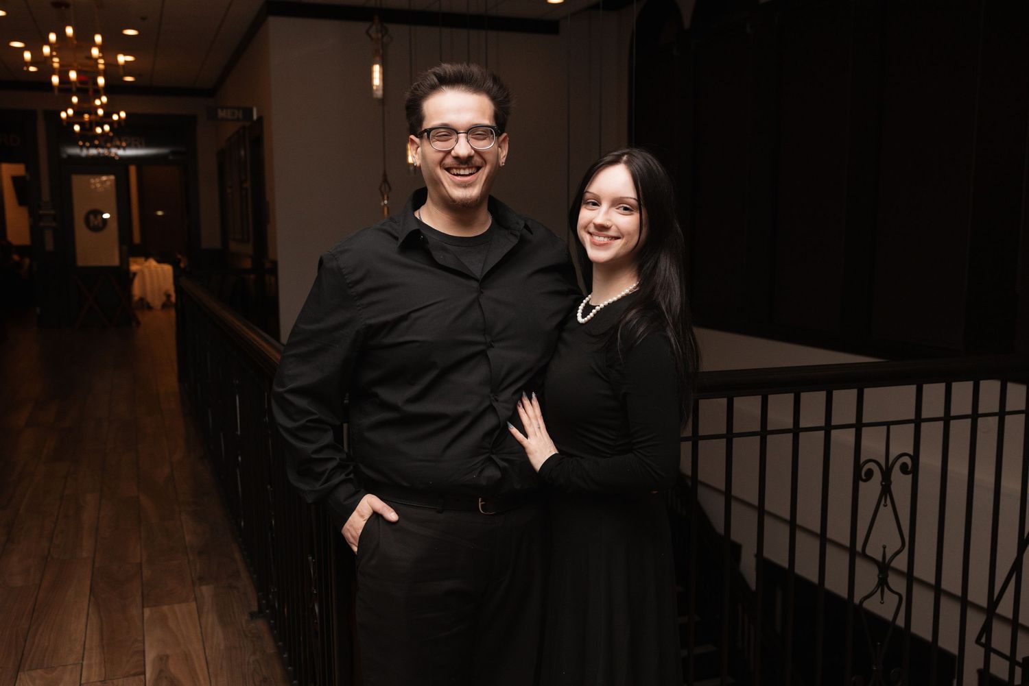 Smiling couple dressed in black posing together in warmly lit venue hallway.