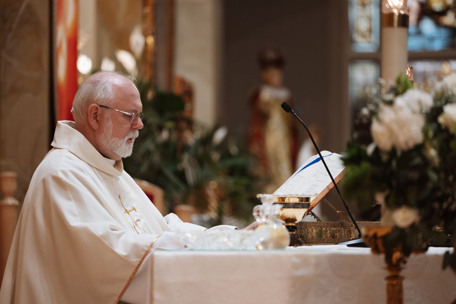 Priest in white vestments reads from book at decorated church altar.