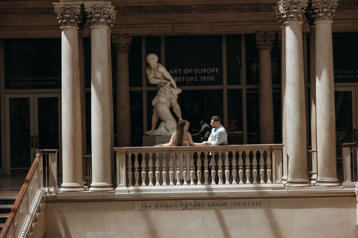 Engaged couple poses on museum balcony beside classical sculpture with grand columns backdrop.