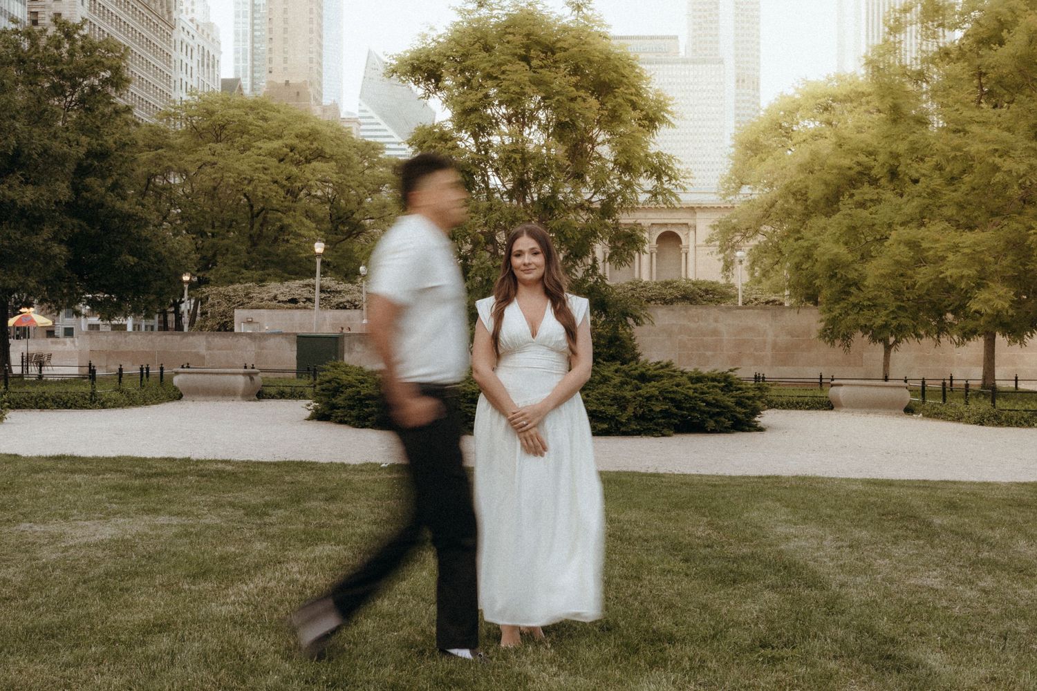 Bride in white gown stands smiling in park as groom walks past in motion blur, city backdrop.