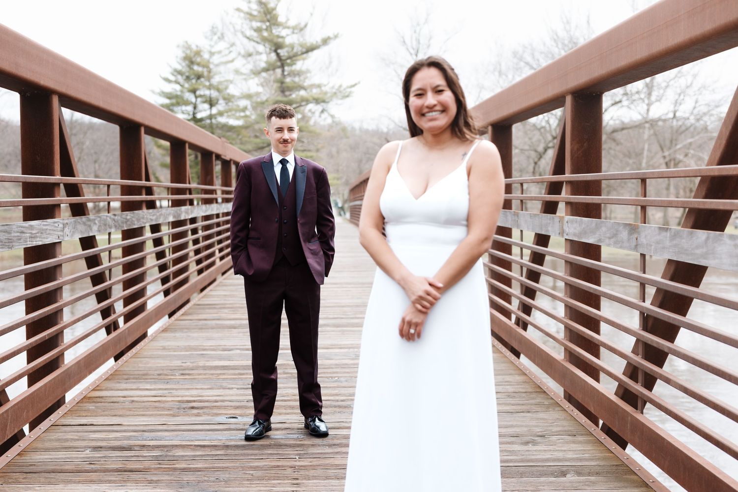 Bride in white gown and groom in burgundy suit pose on a wooden bridge outdoors for wedding photos.