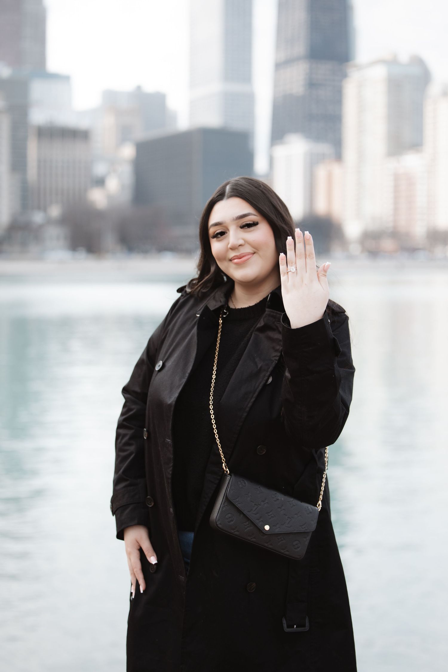 Young woman in black coat waves hand showing engagement ring by Chicago waterfront skyline.