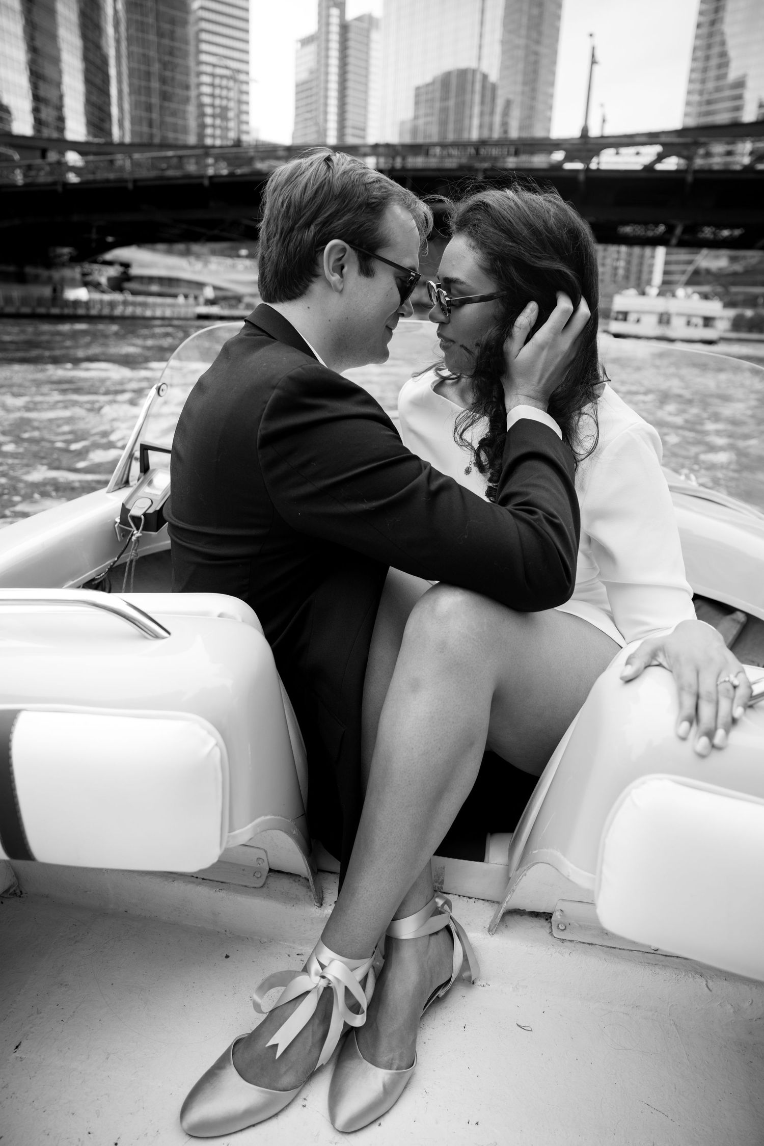 Black and white photo of couple almost kissing on a boat with city skyline behind them.