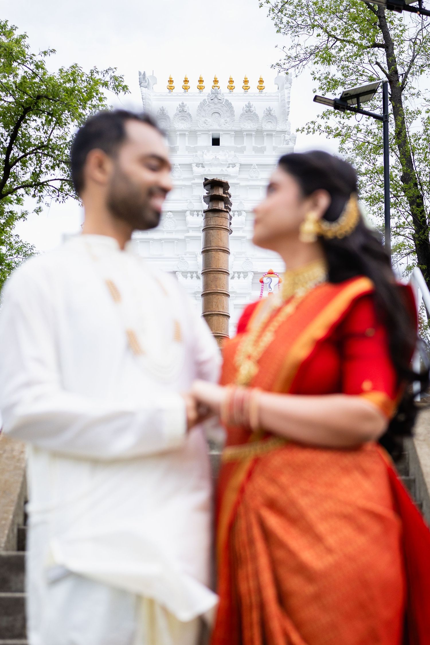 Indian wedding couple in traditional dress standing before white Hindu temple, smiling at each other.
