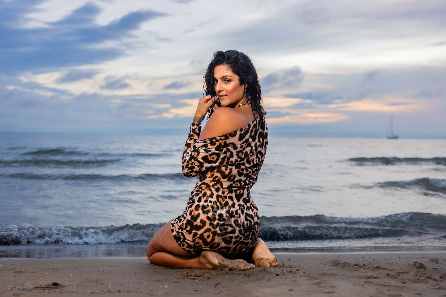 Woman in leopard print dress posing on sandy beach at sunset by sea.