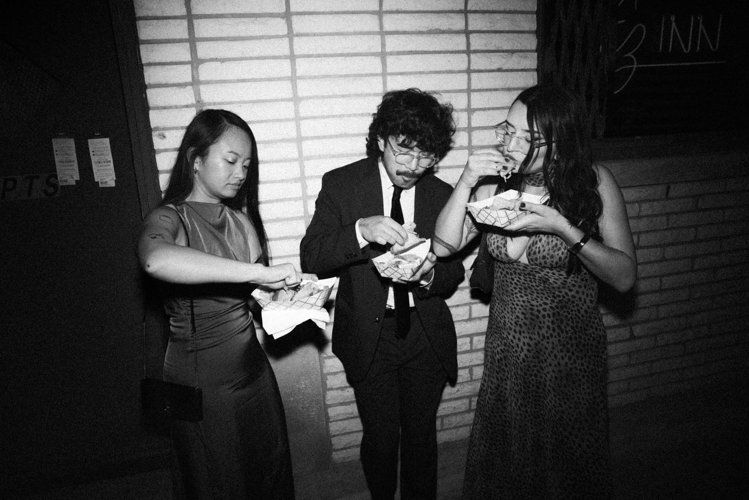 Black and white photo of three formally dressed guests eating food outside a venue at night.