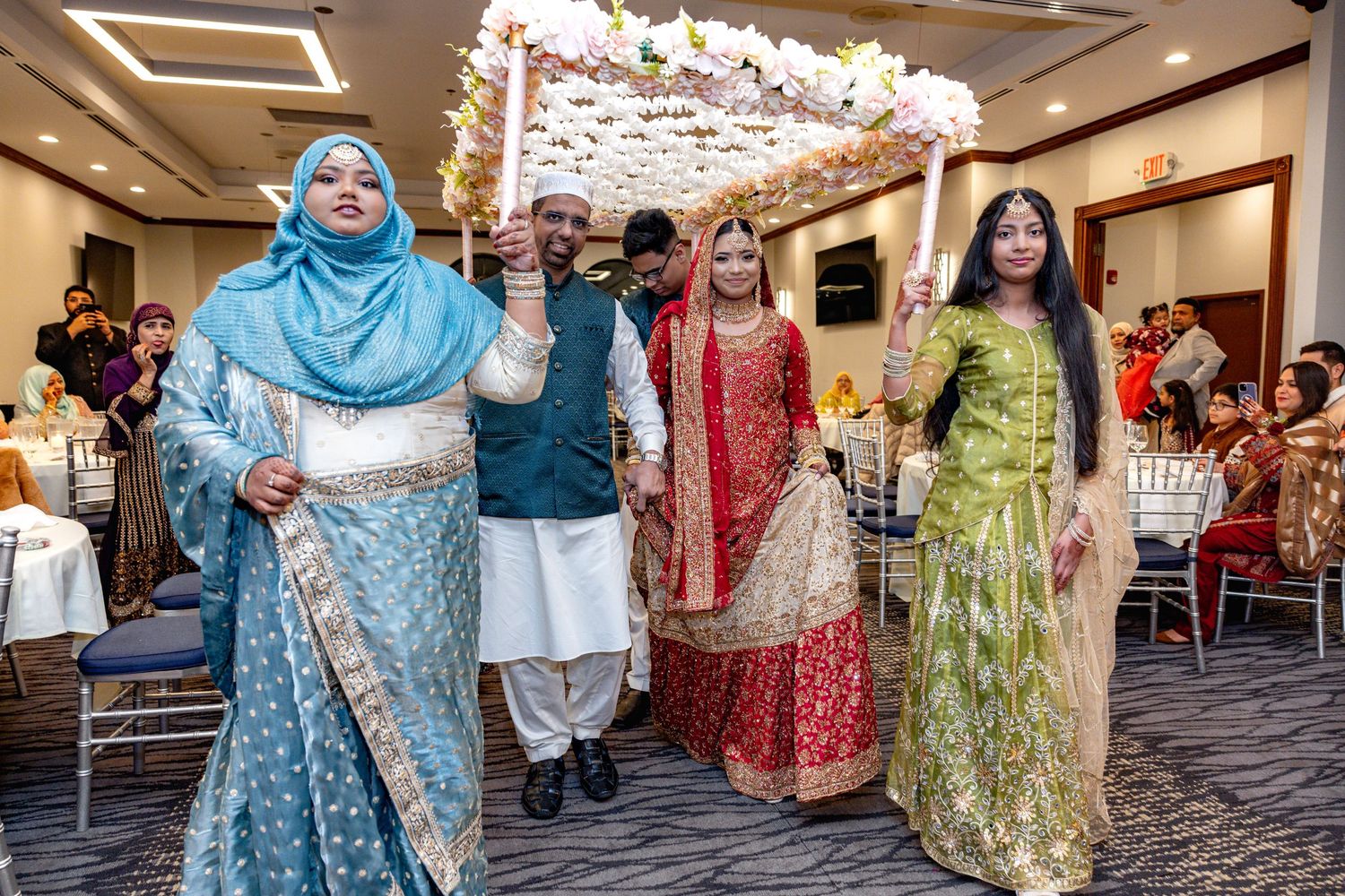 South Asian wedding procession featuring bride under floral canopy with family in traditional clothing.