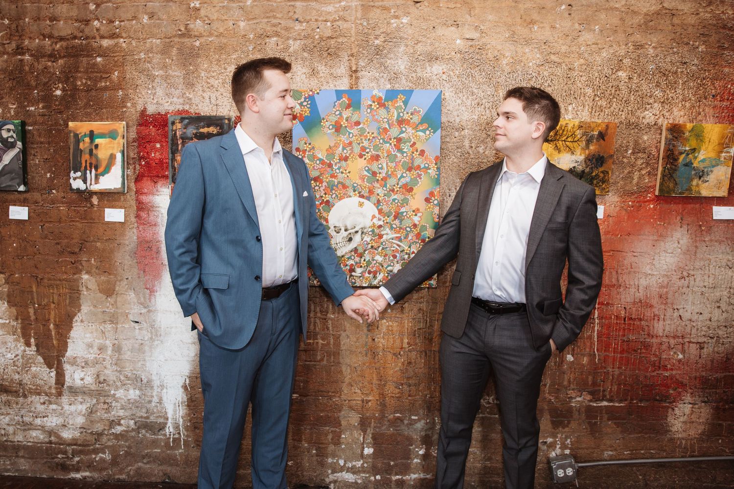 Two men in blue and gray suits sharing a handshake in a rustic art gallery space.