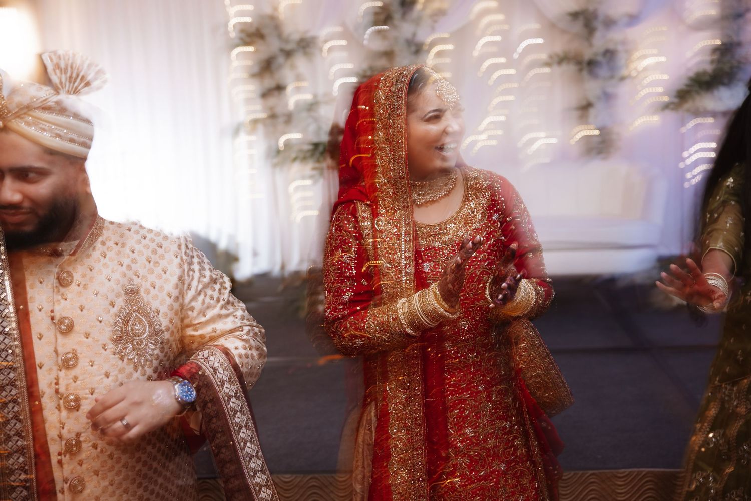 Indian bride smiling in red and gold lehenga with groom in gold sherwani at wedding reception.