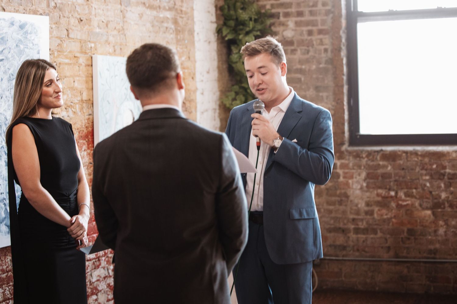 Wedding officiant in blue suit reading vows to couple at elegant indoor brick wall venue.