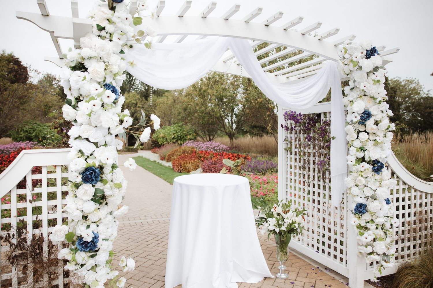 Elegant white wedding arbor adorned with white and navy blue flowers and flowing white drape in garden setting.