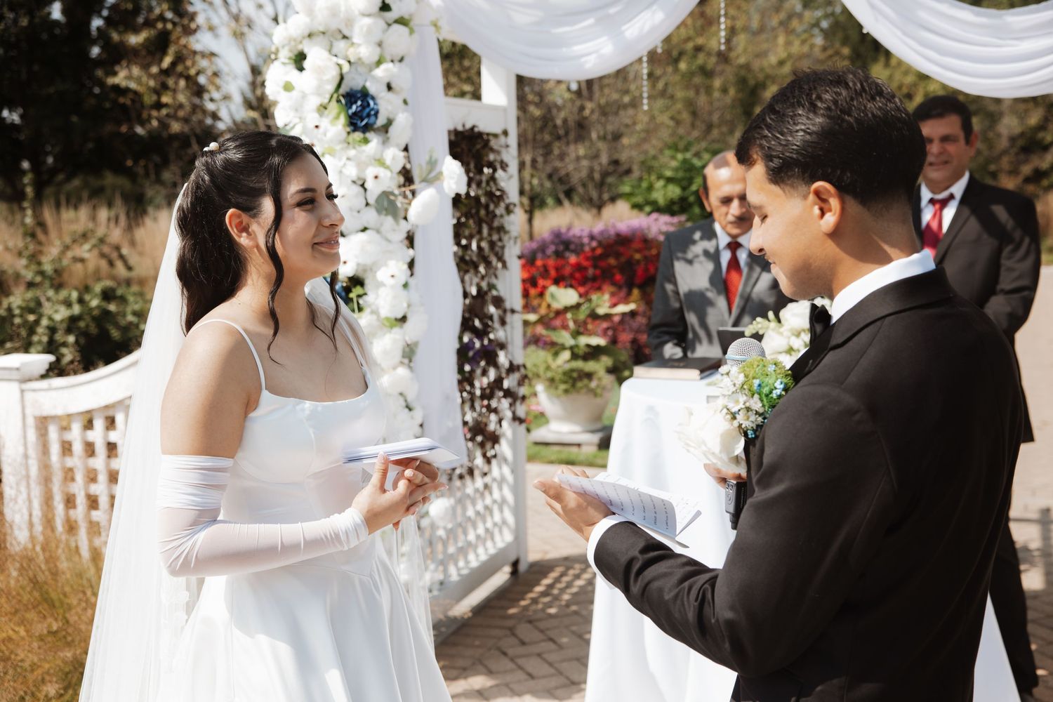 Bride and groom exchanging vows at floral white arch during outdoor wedding ceremony.