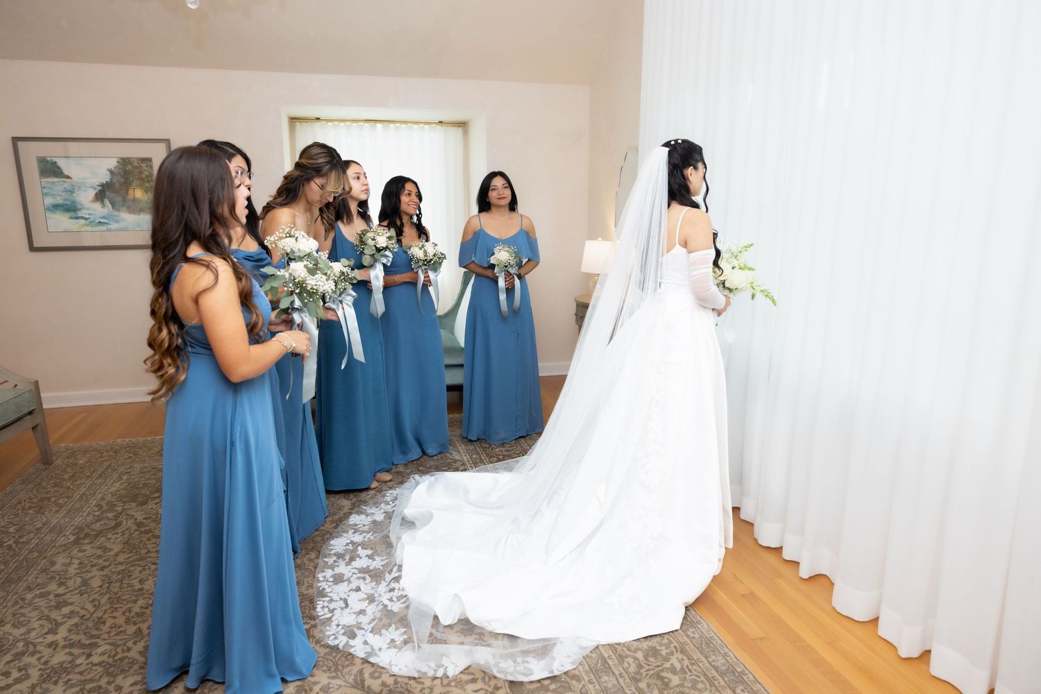 Bridesmaids in steel blue dresses admire bride in lace cathedral veil gown during pre-wedding reveal moment.