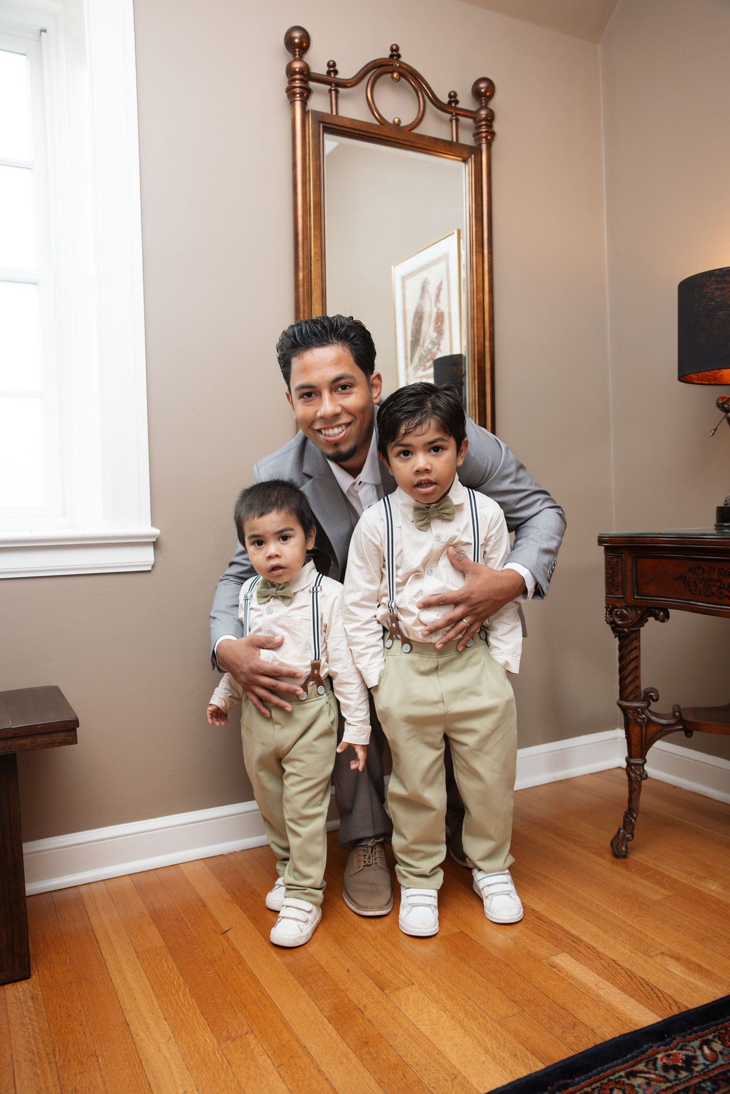Smiling man in gray suit crouching with two young boys in khaki suspenders near decorative mirror.