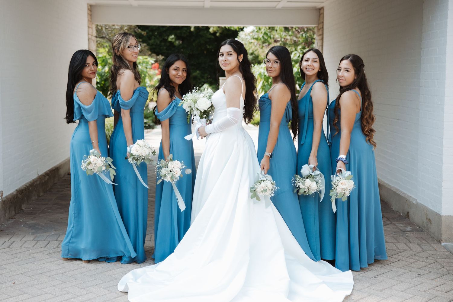 Bride in white gown posed with six bridesmaids in teal dresses holding white bouquets.