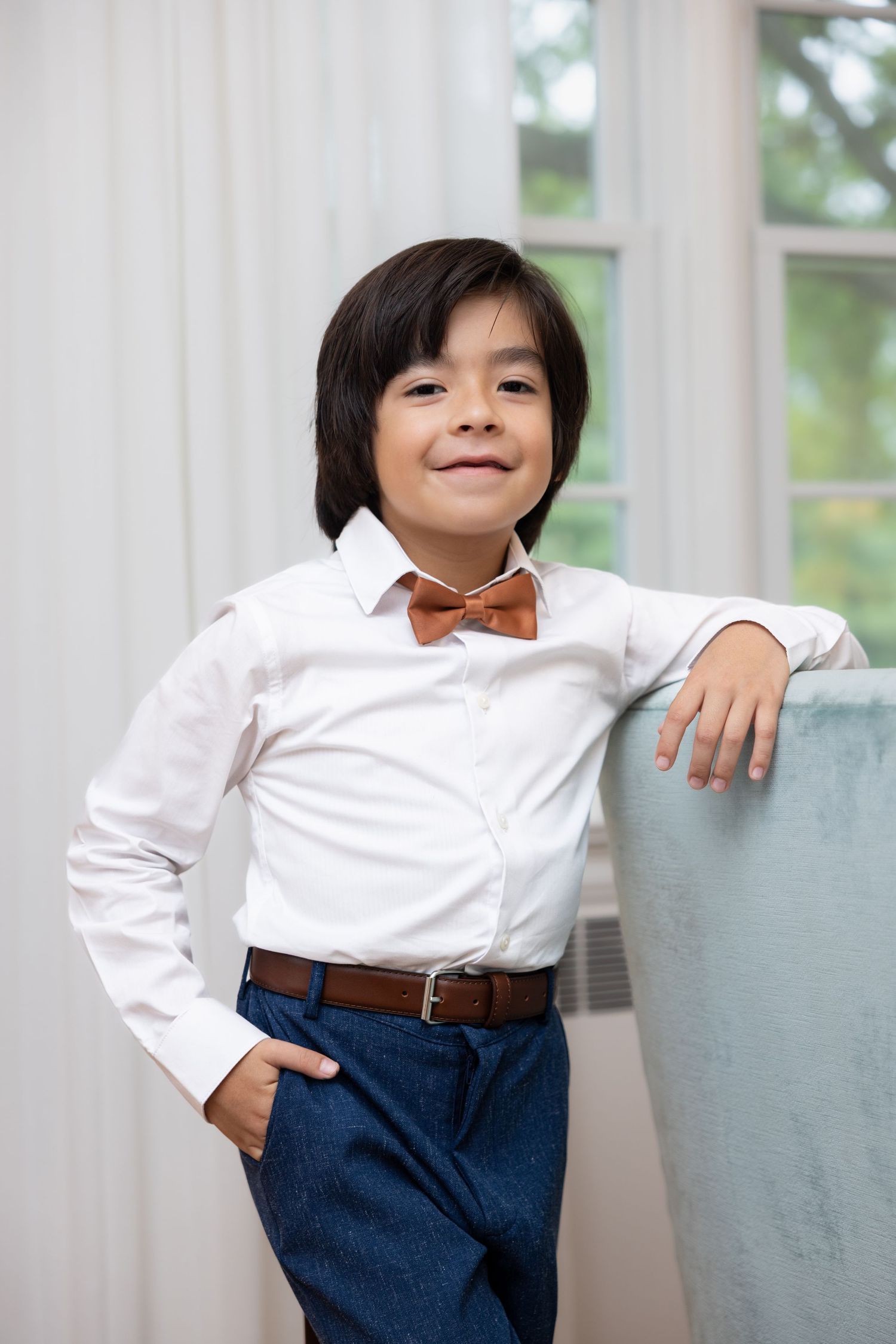 Stylish young boy in white shirt, blue pants, and brown bow tie posing by a sofa.