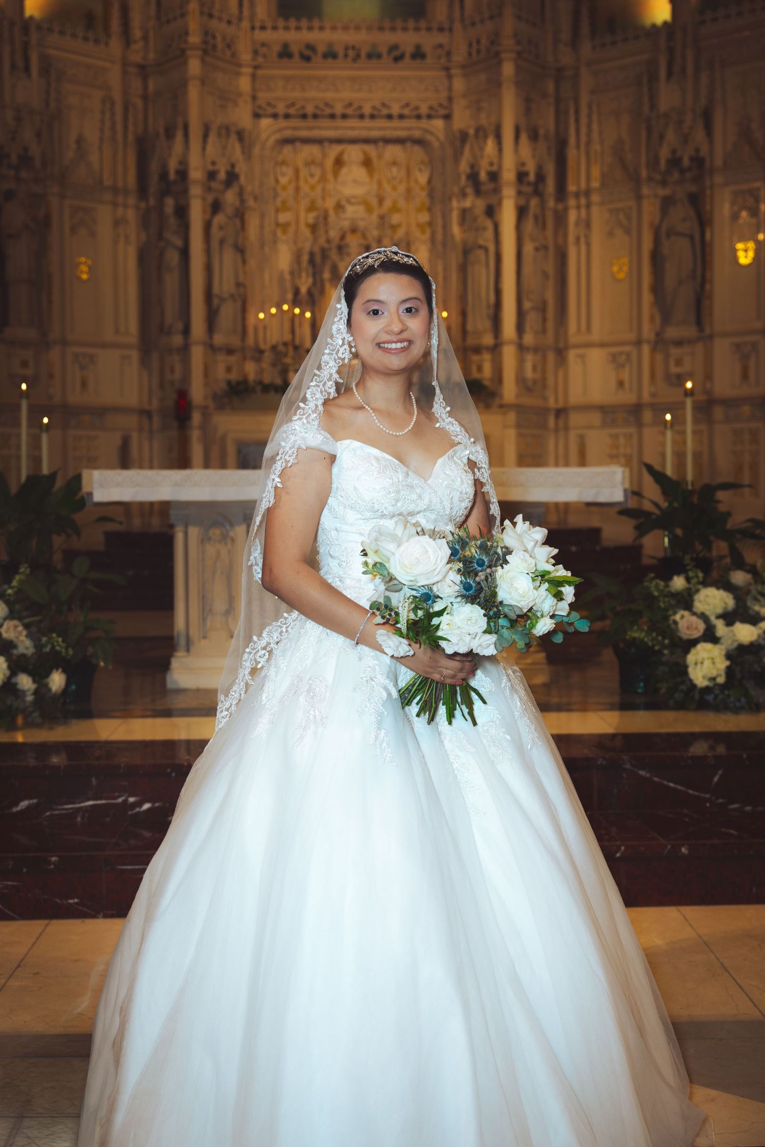 Bride in white ball gown standing at ornate church altar with bouquet.